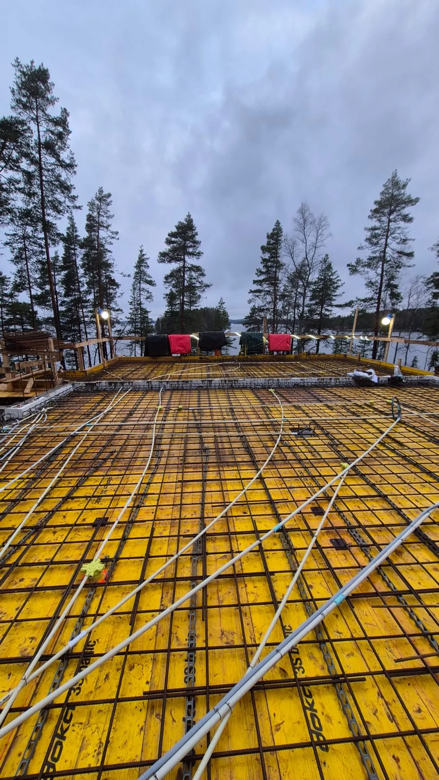 Rakennustyömaa, jossa on keltainen muotti, teräsverkko ja valkoiset putket, taustalla puita ja järvi pilvisellä säällä.