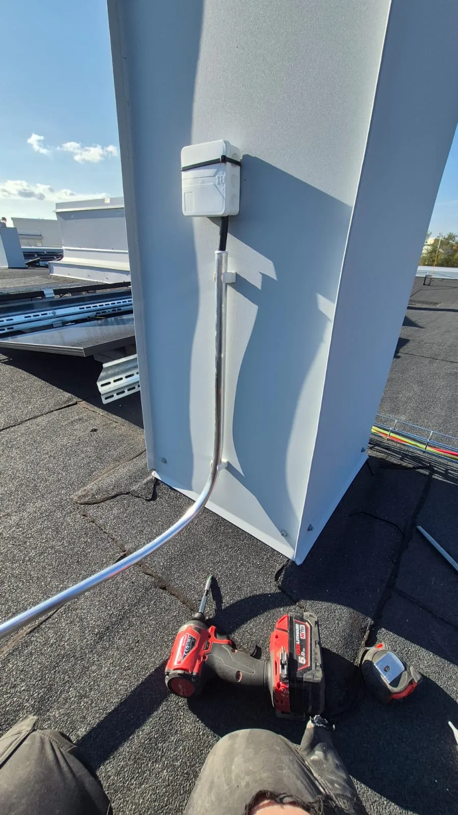 Ulkona otettu kuva, jossa näkyy sähköasennus, mukaan lukien valkoinen kytkentärasia, metalliputki, punainen akkuporakone ja mittanauha mustalla katolla.