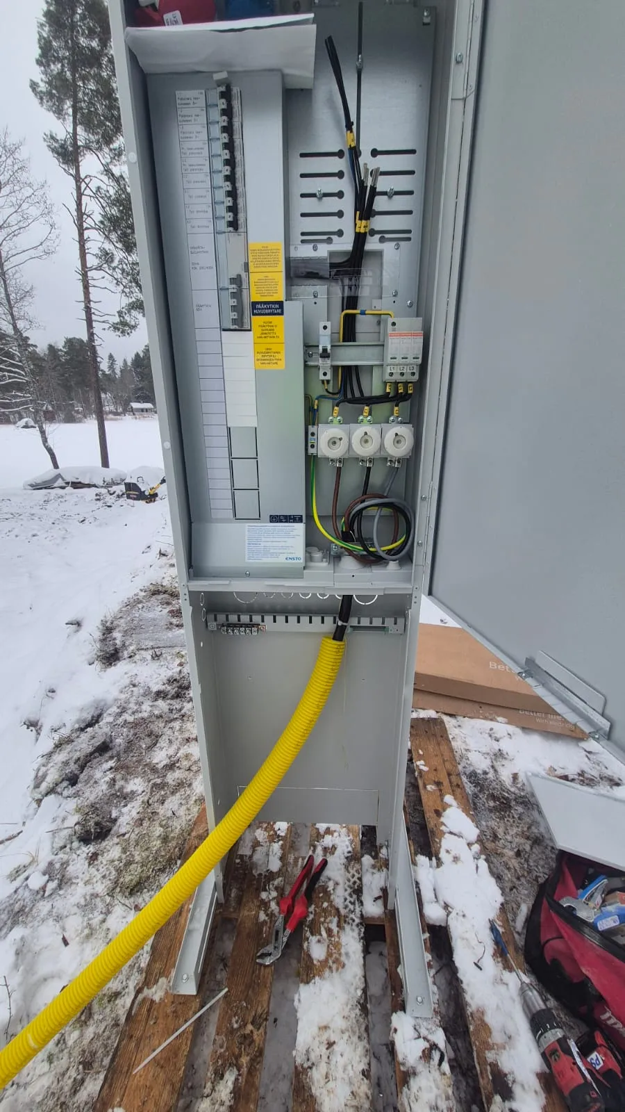 Avoin sähkökaappi lumisessa ulkotilassa, jossa näkyy johdot, sulakkeet ja työkalut. Kirkas valaistus korostaa kaapin sisäosia.