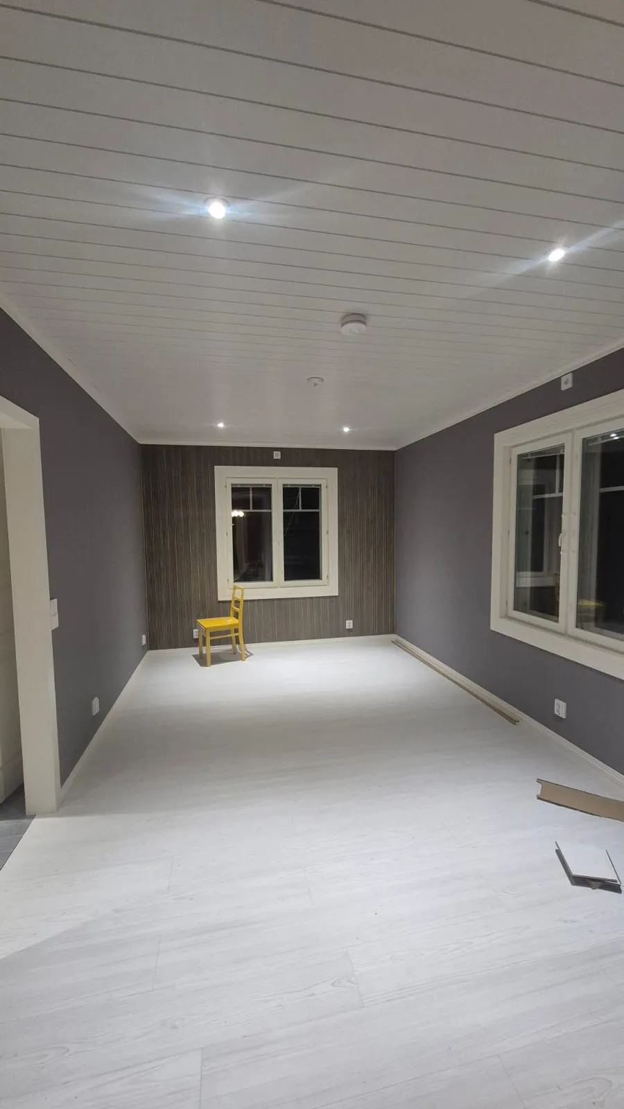 Tyhjä huone, jossa on keltainen tuoli, valkoiset lattiat ja harmaat seinät.