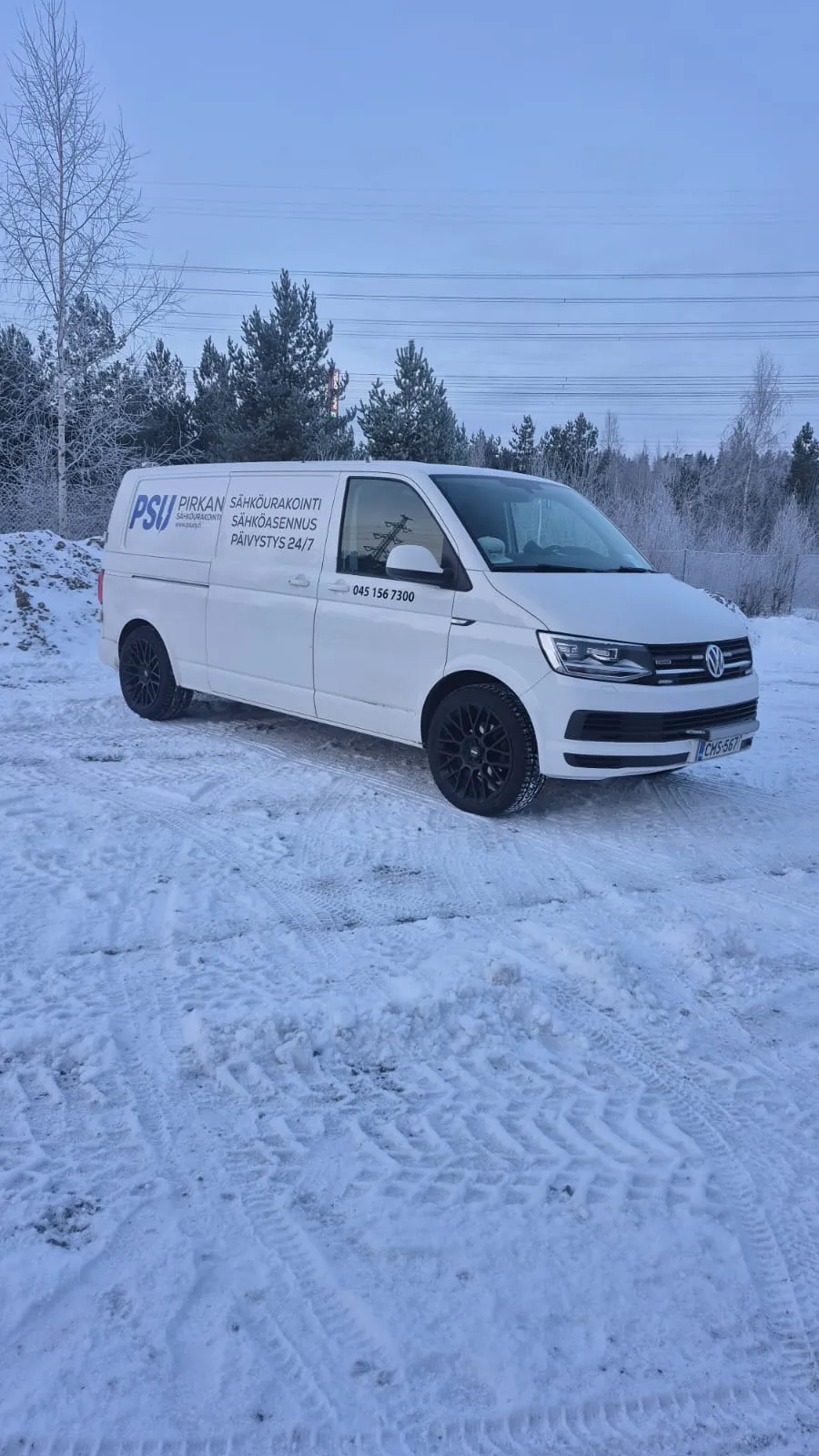 Valkoinen Volkswagen Transporter -pakettiauto lumisella maalla, jossa on PSU Pirkan Sähköurakointi -logo ja puhelinnumero 045 156 7300, taustalla metsä ja talvinen maisema.