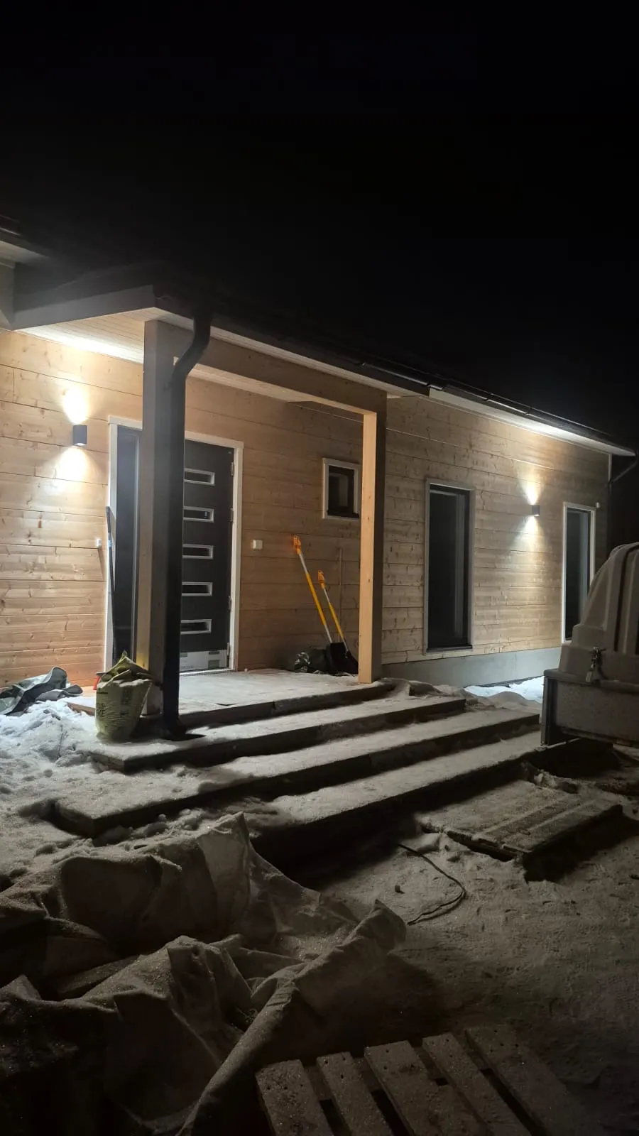 Yöllinen kuva puutalosta, jossa on valaistu kuisti, lumiset portaat ja työkaluja seinustalla.